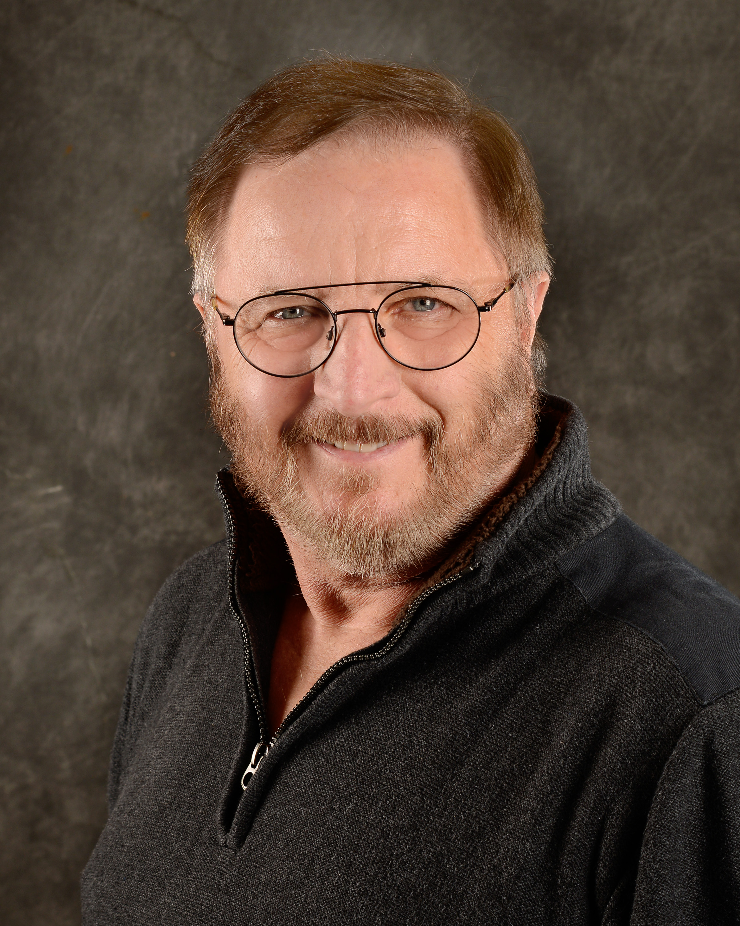 Photo of Bob Gidcumb, dark grey sweater, short brown hair with short boxed beard, smiling wearing wire-rim rounded glasses.