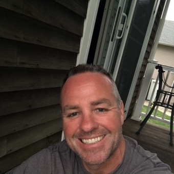 Photo of Brett Legault, in front of a sliding door on a porch or deck, short dark gray hair, heather gray shirt, smiling.