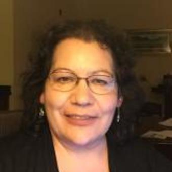 Photo of Carla Osawamick, black shirt, shoulder-length curly dark brown hair, earrings, round-framed glasses, with a subtle smile.