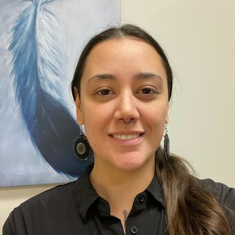 Photo of Chelsey Cameron, painting of an eagle feather on the wall behind, dark brown hair in a pony tail over the left shoulder, round beaded earrings, black dress shirt, septum hoop, smiling.